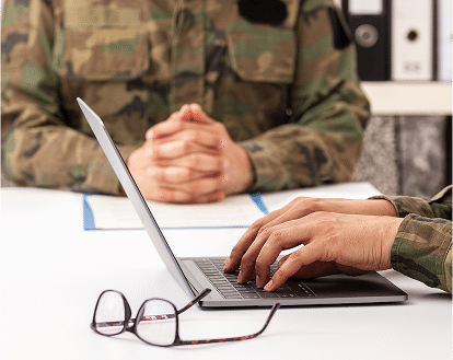 a meeting with two people in camouflage clothing. one with a laptop and glasses on the table.