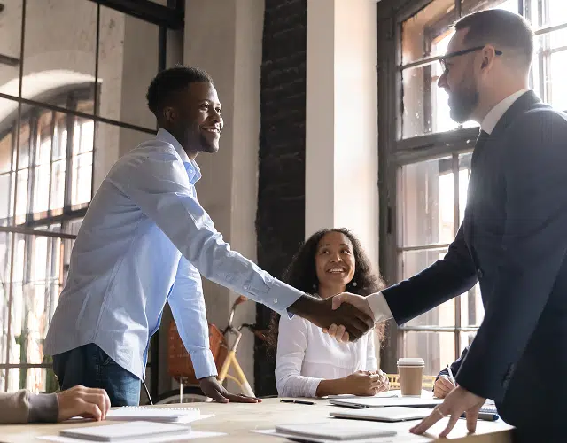 two men standing shaking hands over a table with a woman sitting in the background