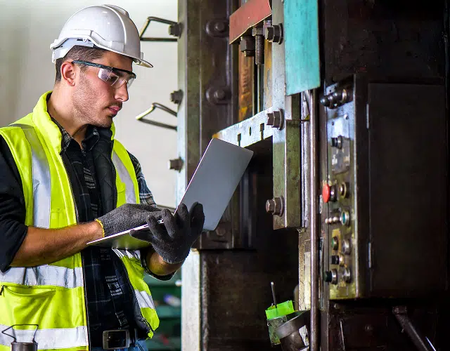 Man wearing a hard hat and high-visibility vest inspecting machinery with a clipboard in hand.