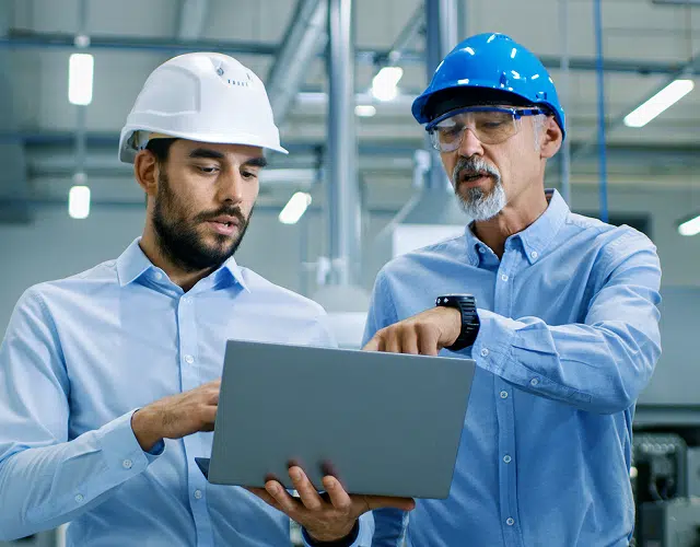 Two workers indoors wearing hard hats and looking at a clipboard